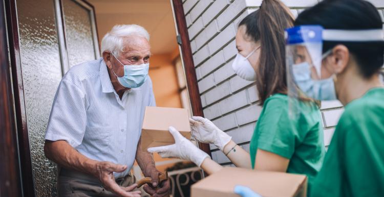 Young female volunteers in mask gives an elderly man boxes with food near his house. Quarantined, isolated. Coronavirus COVID-19. Donation