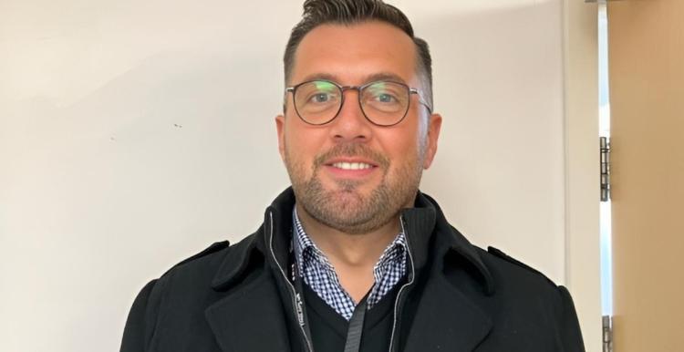 James Treadwell, a medium height man of approx. early 30s, with lightly tanned white skin and short hair styled in a small quiff, smiles warmly facing the camera. He is wearing smart casual outfit of an open-necked checkered shirt with v-neck jumper, both in blue, and a black casual parka. He is wearing brown framed glasses and a black lanyard carrying a brightly coloured ID badge. He is photographed against a cream background similar to a public building interior wall.