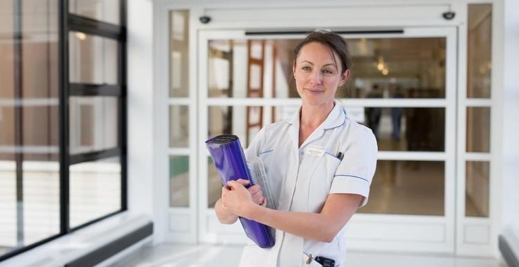 A nurse in a hospital corridor holding a folder and smiling